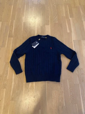 Mörkblå kabelstickad tröja Polo Ralph Lauren - Mörkblå kabelstickad tröja från Polo Ralph Lauren med klassisk rund halsringning och röd broderad logga på bröstet. Tröjan har ribbade muddar vid ärmslut och nederkant. Tillverkad i mjukt material som ger en skön och stilren look.