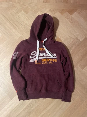 Vinröd hoodie från Superdry - Snygg vintage vinröd hoodie från Superdry med vit och gul text på bröstet samt japanska tecken. Bra kvalité och perfekt vintage. Hör av er vid funderingar.