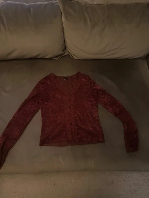 Vinröd spetskofta med knappar - Snygg vinröd kofta i spetsmaterial med långa ärmar och knäppning framtill. Kortare modell med v-ringning och diskret blommönster i tyget. Perfekt för att lyfta en outfit med lite extra detaljer.