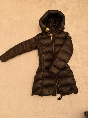 Svart lång dunjacka med huva - Snygg svart lång dunjacka från parajumper med huva och dragkedja framtill. Jackan är quiltad och har en figurnära passform som håller dig varm under kalla dagar. Perfekt för vintern och enkel att styla till olika outfits.