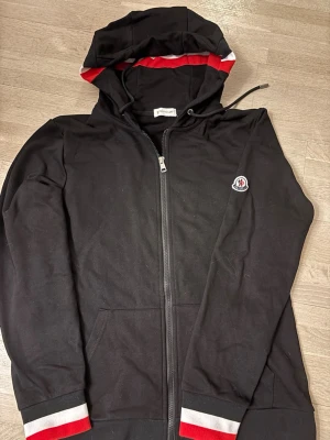 Svart Moncler hoodie med dragkedja - Snygg svart hoodie från Moncler med dragkedja framtill och huva med dragsko. Röd och vit detalj på huva och ärmslut samt Moncler-logga på ärmen. Tillverkad i mjukt bomullsmaterial, perfekt för en avslappnad och trendig look.
