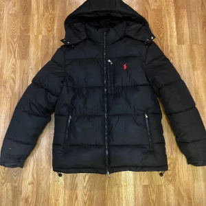 Svart dunjacka från Ralph Lauren - Svart puffer dunjacka från Ralph Lauren med huva och dragkedja framtill. Jackan har två snedställda fickor med dragkedja och en liten röd logga broderad på bröstet. Perfekt för kalla dagar och har en klassisk, sportig look.