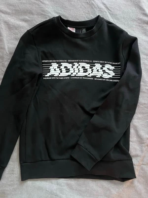 Svart sweatshirt från Adidas - Säljer en svart sweatshirt från Adidas med stor vit logga och text på bröstet. Tröjan har rund halsringning, långa ärmar och ribbade muddar vid ärmslut och nederkant. Perfekt för en sportig och avslappnad stil.