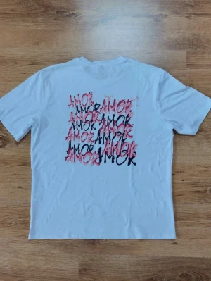 Vit t-shirt med färgglad text - Vit t-shirt med trycket 'AMOR' i flera färger på ryggen. T-shirten har rund hals och korta ärmar. Materialet känns som bomull och passformen är normal. Perfekt för dig som gillar grafiska tryck och enkel stil.
