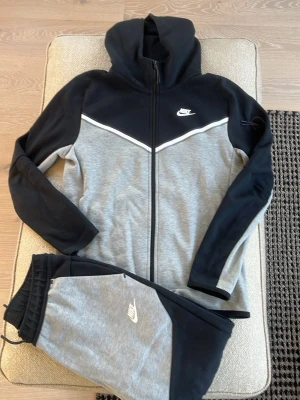 Nike grå och svart tracksuit set - Nike tracksuit i tvådelad design med huvtröja och byxor. Färgerna är grå och svart med vit Nike-logga på både tröja och byxa. Tröjan har dragkedja och huva, byxorna har resår i midjan och muddar vid benslut. Perfekt för chill eller träning.