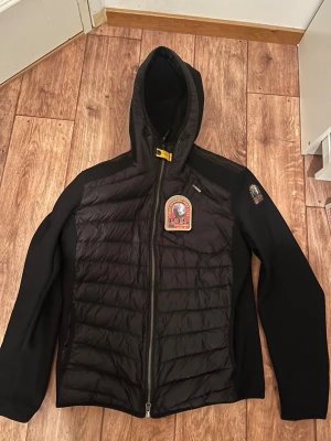Svart pufferjacka med huva från Parajumpers - Svart pufferjacka från Parajumpers med huva och dragkedja framtill. Jackan har quiltade partier på bröst och rygg, samt märkespatch på bröstet och ärmen. Tillverkad i polyester och syntetmaterial för en sportig look. Priset går att diskuteras vid snabb affär 