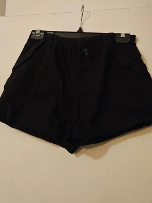 Svarta shorts med snörning - Svarta shorts med elastisk midja och snörning framtill. Modellen har två sidofickor och en låtsasbakficka med lock. Perfekta för tex festival!