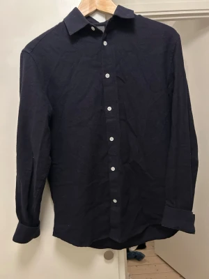 Svart skjorta med knappar - Stilren svart skjorta med klassisk krage och vita knappar framtill. Skjortan har lång ärm och är tillverkad i ett mjukt tyg som känns bekvämt mot huden. Perfekt för dig som gillar en clean och enkel look.