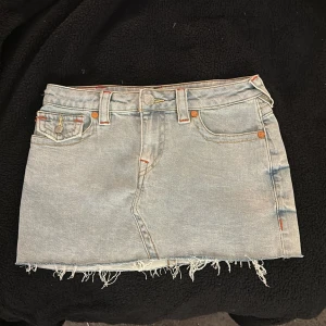 True Religion Sadie jeanskjol ljusblå - Ljusblå jeanskjol från True Religion, modell Sadie Mini Skirt. Kjolen har rå, fransig nederkant, klassiska fickor fram och bak med färgglada broderier och logoknappar. 