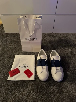 Valentino Garavani Open  - Säljer ett par snygga Valentino Garavani Open sneakers i vitt skinn med bred blå rem över vristen och ikoniska nitar på hälen. Klassisk rund tå och platt sula. Kommer med originalpåse och förpackning.