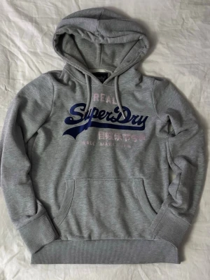 Grå Superdry hoodie - Snygg grå hoodie från Superdry med stor logga i blått och rosa framtill. Klassisk känguruficka, ribbade muddar och huva med dragsko. Tillverkad i mjukt bomullsmaterial som är perfekt för chill dagar. Japansk text under loggan ger extra streetkänsla.