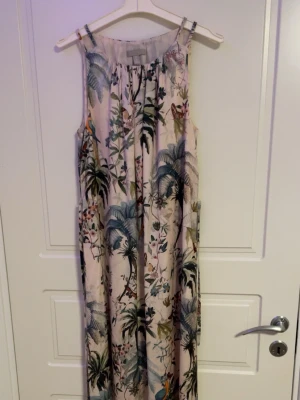 Långklänning med tropiskt mönster - Säljer en ärmlös långklänning med tropiskt blommigt mönster i grönt, blått och beige. Klänningen har rund halsringning och ett luftigt, flowy fall. Perfekt för sommardagar och semester. Materialet känns lätt och svalt, troligen polyester eller bomull.