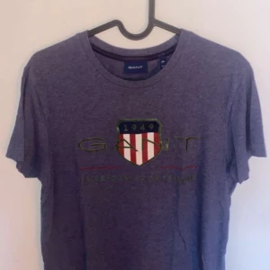 Blå t-shirt från GANT - Snygg blå t-shirt från GANT med broderad logga och sköld på bröstet. Klassisk rund halsringning och korta ärmar. Perfekt för en avslappnad och sportig stil.