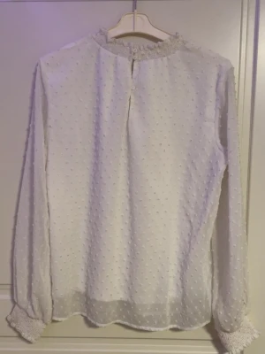 Vit prickig blus med smockdetaljer - Säljer en vit blus med diskreta prickar i tyget och smock vid halsringning och ärmslut. Blusen har en lös passform och är tillverkad i ett tunt, transparent material som ger en luftig känsla. Perfekt att styla med jeans eller kjol för en trendig look.