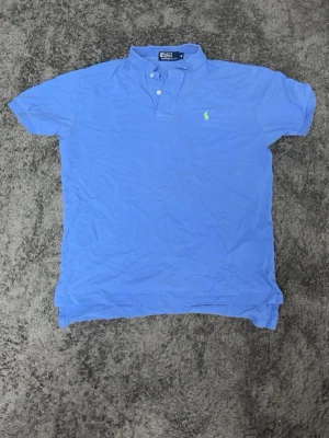 Polo Ralph Lauren piké  - Klassisk blå pikétröja från Polo Ralph Lauren med korta ärmar och vit knappslå. Tröjan har den ikoniska broderade loggan i grönt på bröstet och är tillverkad i bomull för en bekväm känsla. Perfekt till jeans eller shorts.