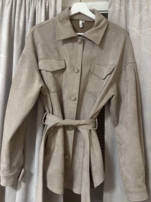 Beige overshirt med bälte från Nelly - Stilren beige overshirt från Nelly i mjukt mocka material med knappar framtill, två bröstfickor med lock och ett matchande bälte i midjan. Jackan har klassisk krage och långa ärmar med knappdetalj vid mudden. Perfekt för lager på lager och en clean look.