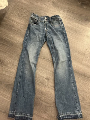 Blå jeansbyxor från Gina Tricot - Säljer ett par klassiska blå jeansbyxor från Gina Tricot med raka ben och rå kant nertill. Jeansen har normal passform, fem fickor och snygga slitningar framtill. Materialet är slitstark denim i bomull och de har dubbla knappar i midjan.