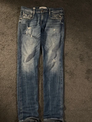 Replay jeans slitningar - Säljer ett par blå jeans från Replay med slitna detaljer och slimmade ben. Jeansen har klassisk femficksdesign, normal passform och är tillverkade i bomull med en snygg tvättad look. Perfekta för dig som gillar en avslappnad stil. W29 l 32
