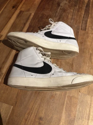 Nike Blazer Mid vita sneakers - Klassiska Nike Blazer Mid sneakers i vitt skinn med svart swoosh på sidan. Högt skaft, snörning och retroinspirerad design. Tidlös modell som passar till det mesta och har en platt sula samt rund tå.