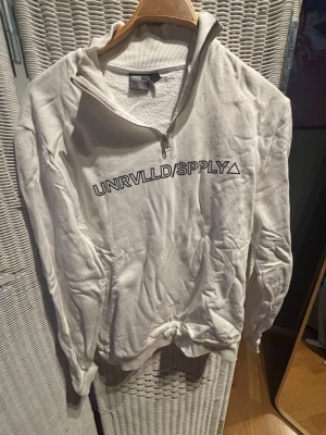 Vit quarter zip hoodie UNRVLLD/SPPLY - Säljer en vit hoodie från UNRVLLD/SPPLY med quarter zip och hög krage. Hoodien har tryckt logga på bröstet och är gjord i mjukt sweatshirtmaterial. Perfekt för en avslappnad och trendig look.