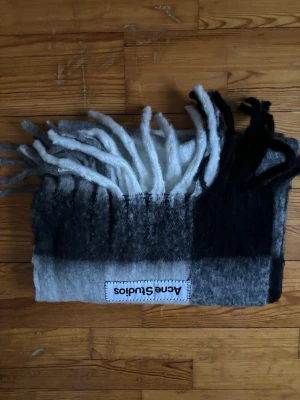 Rutig halsduk från Acne Studios - Mjuk och fluffig halsduk från Acne Studios i grå, svart och vit ton. Halsduken har ett rutigt mönster och långa fransar i ändarna. Perfekt accessoar för att lyfta din outfit med en stilren touch.