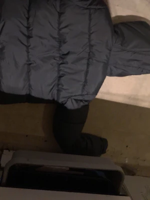 Mörkgrå puffer dunjacka med huva - Snygg grå/svart dunjacka i puffer-modell med huva och dragkedja framtill. Jackan har långa ärmar och quiltad design som ger en riktigt modern look. Perfekt för kalla dagar när du vill hålla dig varm och stilren.