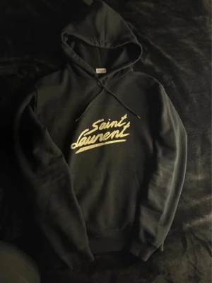 Svart Saint Laurent hoodie - Snygg svart hoodie från Saint Laurent med vit logga i retrostil på bröstet. Klassisk huva med dragsko och stor magficka framtill. Tillverkad i mjukt material som känns skönt mot huden. Perfekt för dig som vill ha en stilren och trendig look.