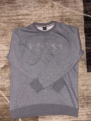 Grå sweatshirt från Hugo Boss - Säljer en stilren grå sweatshirt från Hugo Boss med stor logga framtill. Tröjan har rund hals, ribbade muddar och är tillverkad i mjuk bomullsmix. Perfekt för dig som gillar enkel och clean stil.