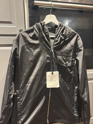 Svart vindjacka från Moncler - Snygg svart vindjacka från Moncler med huva och dragkedja framtill. Jackan har en bröstficka med knapp och minimalistisk design. Tillverkad i "smart cashmere" som passar perfekt för blåsiga dagar. Klassisk passform och stilren look. Fick i julklapp behöver pengar till bil, har inte kvitto