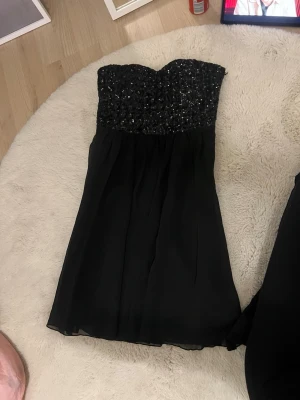 Svart axelbandslös paljettklänning - Snygg svart kortklänning med axelbandslös design och glittrig paljettöverdel. Kjolen är i ett lätt, flygigt tyg som ger en fin siluett. (den är inte genomskinlig) den är knappt använd.