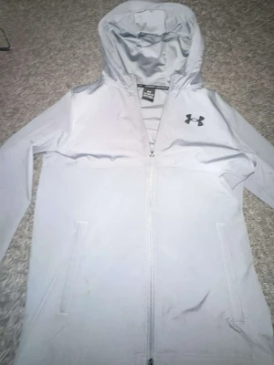 Ljusgrå vindjacka från Under Armour - Snygg ljusgrå vindjacka från Under Armour med huva och dragkedja framtill. Jackan har två sidofickor och en diskret logga på bröstet. Tillverkad i lätt och smidigt material som passar perfekt för blåsiga dagar.