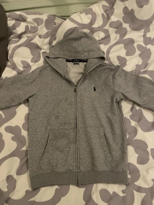Grå hoodie från Polo Ralph Lauren - Klassisk grå hoodie från Polo Ralph Lauren med dragkedja framtill och huva. Tröjan har två fickor fram och den ikoniska lilla loggan broderad på bröstet. Tillverkad i mjuk bomull så är väldigt skön. Strrlek M men passar perfekt som S, finns inga defekter. Kom med frågor o prisförslag