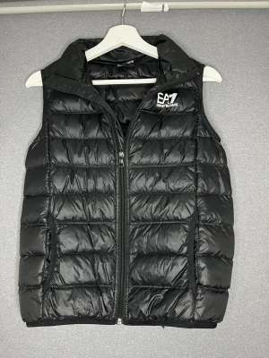 Svart dunväst från EA7 Emporio Armani - Snygg svart dunväst från EA7 Emporio Armani med quiltad design och dragkedja framtill. Västen har hög krage och två fickor med dragkedja. Perfekt för lager-på-lager och ger en sportig look. Märkeslogga i vitt på bröstet för extra stilpoäng.