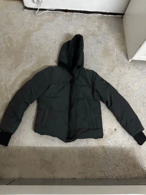 Mörkgrön pufferjacka från Canada Goose Macmillian black label - Säljer en svart pufferjacka från Canada Goose med huva och diskret logga på ärmen. Jackan har stora fickor framtill, dragkedja och knappar. Materialet är polyester och bomull på utsidan, fodrad med dun och fjädrar för extra värme. Perfekt för kalla dagar.