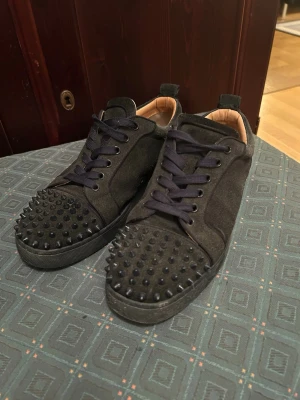 Christian Louboutin  - Säljer ett par mörkblåa sneakers i mocka från Christian Louboutin med coola svarta nitar på tån och klassisk röd sula. Skorna har svarta snören och en låg profil. Insidan är beige och materialet känns lyxigt och mjukt. En nit saknas men syns inte 