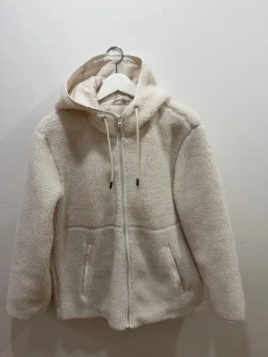 Vit teddyjacka med huva och dragkedja - Mysig vit teddyjacka med huva och dragsko. Jackan har hel dragkedja framtill och två fickor med dragkedja. Perfekt för kyliga dagar när du vill vara både varm och stilren. 