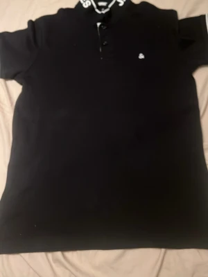 Svart piké från Jack & Jones storlek:S - Snygg svart pikétröja från Jack & Jones med vit logga på bröstet och textdetaljer på kragen. Tröjan har korta ärmar och knappar vid halsen. Perfekt för en clean och sportig stil.