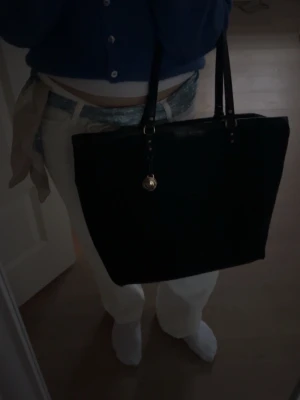 Svart shoppingväska från Tommy Hilfiger - Snygg svart shoppingväska från Tommy Hilfiger med diskret mönster och guldfärgad logodetalj. Väskan har två långa handtag och är perfekt för att bära allt du behöver. Materialet är en mix av textil och syntet med läderdetaljer. Perfekt för dig som vill ha både stil och funktion. Får plats med MacBook och nästan oanvänd!