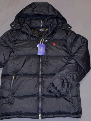 Svart dunjacka från Polo Ralph Lauren - Snygg svart dunjacka från Polo Ralph Lauren med avtagbar huva och dragkedja framtill. Jackan har två sidofickor med dragkedja och den klassiska röda loggan broderad på bröstet. Perfekt för kalla dagar och riktigt stilren.