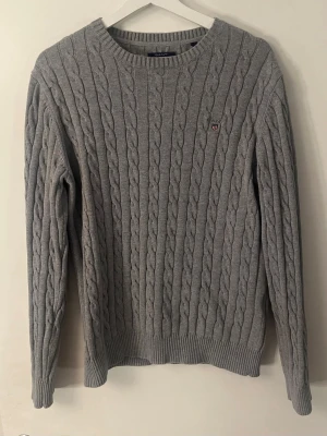 Grå kabelstickad tröja från Gant - Klassisk grå kabelstickad tröja från Gant L men passar M med rund halsringning och diskret logga på bröstet. Tröjan har långa ärmar och ribbade muddar vid ärmslut och nederkant och pris kan diskuteras vid snabb affär