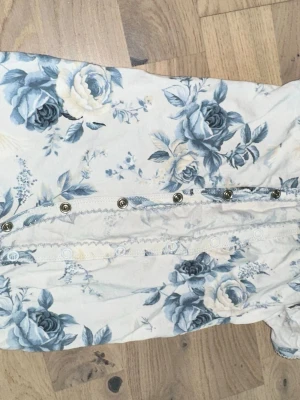 Blommig body från Newbie, stl 68 - Supersöt body från Newbie i storlek 68 med blått och vitt blommönster. Bodyn har tryckknappar framtill och vid grenen samt fina spetsdetaljer längs kanterna. Tillverkad i mjuk bomull, perfekt för små barn.