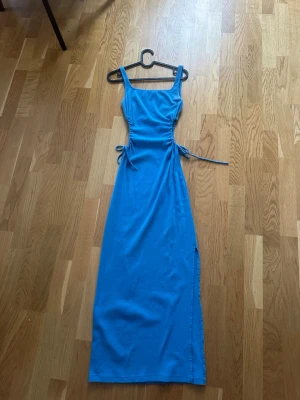 Blå långklänning med snörning - Snygg blå långklänning med smala axelband och snörning i midjan på båda sidor. Klänningen har en rak passform och slits på sidan, perfekt för en trendig look. Materialet känns mjukt och stretchigt.