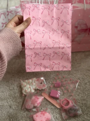 Rosa presentpåse med rosa assesories - Söt rosa presentpåse med tryckta rosett-mönster och ett handtag i vitt snöre. Påsen har en stor satinrosett framtill som extra detalj. Perfekt för att ge bort små presenter eller accessoarer. Liten och smidig storlek, supergullig design.great idea for gift like birthdays and valentines gift