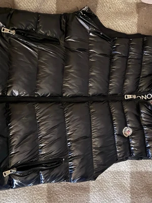 Svart dunväst från Moncler L - Snygg svart dunväst från Moncler med glansig finish och klassisk quiltad design. Västen har två dragkedjeförsedda fickor framtill och Moncler-logga på bröstet. Perfekt för lager-på-lager och streetwear-stil.