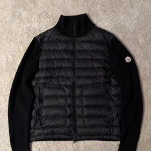 Moncler Cardigan - Snygg svart dunväst från Moncler med quiltad front och två dragkedjefickor. Västen har hög krage och Moncler-logga på ärmen. Perfekt för lager-på-lager och ger en clean look till din outfit. Materialet är en mix av dun och textil för extra komfort. Står storlek XL, men passar M/L - Cardiganen är lätt använd, inga stora tecken på användning