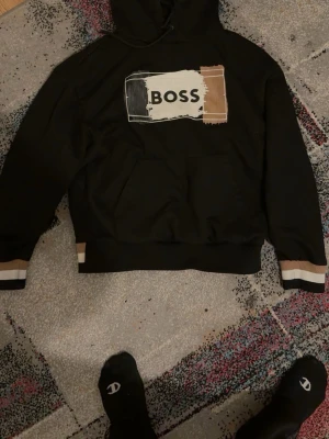 Svart hoodie från BOSS med patch - Svart hoodie från BOSS med stor patch framtill i svart, vitt och beige med texten 'BOSS'. Huva med dragsko och känguruficka. Muddar och nederkant har beige och vita detaljer. Perfekt för en avslappnad streetwear-look.