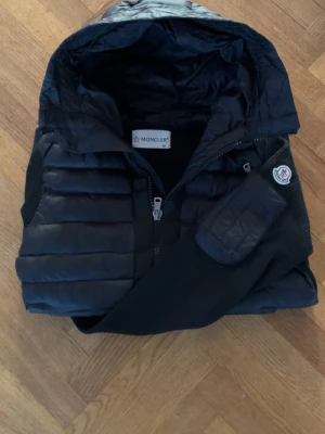 Svart dunväst från Moncler med huva - Snygg svart dunväst från Moncler med huva och quiltad design. Västen har dragkedja framtill, Moncler-logga på axeln och praktiska fickor. Perfekt för lager-på-lager och streetwear-stil.