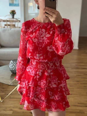 Röd blommig volangklänning - Säljer en röd kortklänning med rosa blommönster och volanger. Klänningen har långärmade, transparenta ärmar med resår vid handlederna och smock i midjan för en snygg passform.