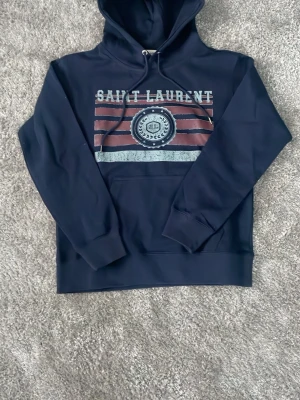 Mörkblå Saint Laurent hoodie - Snygg mörkblå hoodie från Saint Laurent med tryck framtill i vitt, rött och grått. Klassisk huva med dragsko och ribbade muddar vid ärmar och nederkant. Perfekt för dig som vill ha en stilren och trendig look.