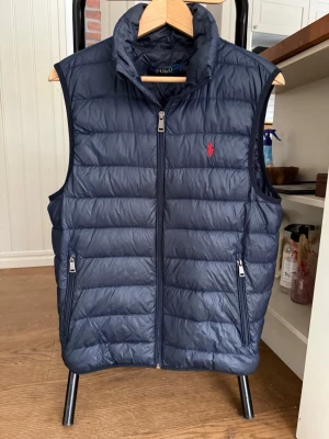 Mörkblå dunväst från Polo Ralph Lauren - Snygg mörkblå dunväst från Polo Ralph Lauren med hög krage och dragkedja framtill. Västen har två dragkedjeförsedda fickor och den klassiska röda loggan broderad på bröstet. Perfekt för lager-på-lager och streetstyle.